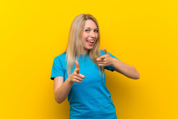 Young blonde woman over isolated yellow background pointing to the front and smiling