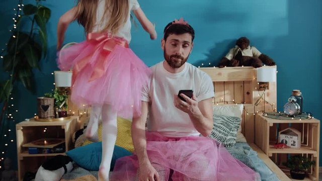 Portrait of exhausted young parent father using a smartphone sitting on bed and touching his face. Handsome young man is tired of playing with kids. Fancy cute girl dancing and having fun on bed