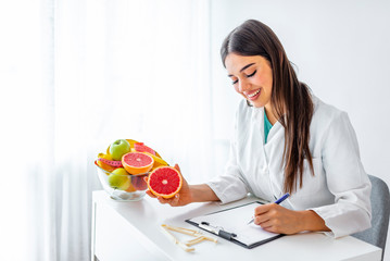 Obraz na płótnie Canvas Portrait of young smiling female nutritionist in the consultation room. Nutritionist desk with healthy fruit, juice and measuring tape. Dietitian working on diet plan.