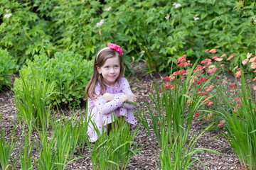 Horizontal full length view of pretty fair small girl crouching in flowerbed wearing a mauve summer...