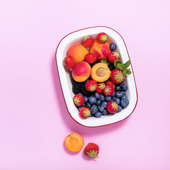 Ripe apricots and summer berries in a bowl on pink paper background square image