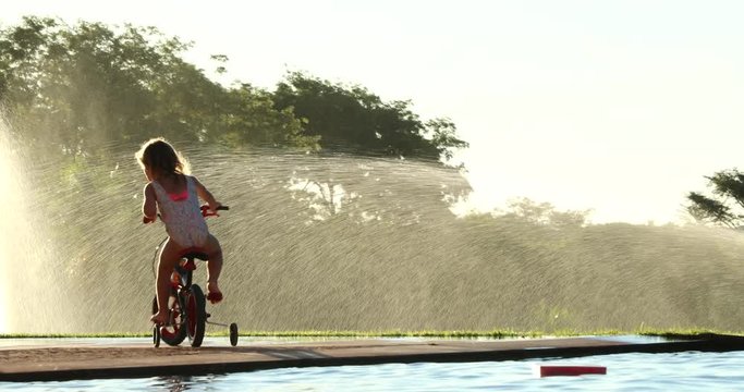Little girl riding tricycle with water sprinkles in the background