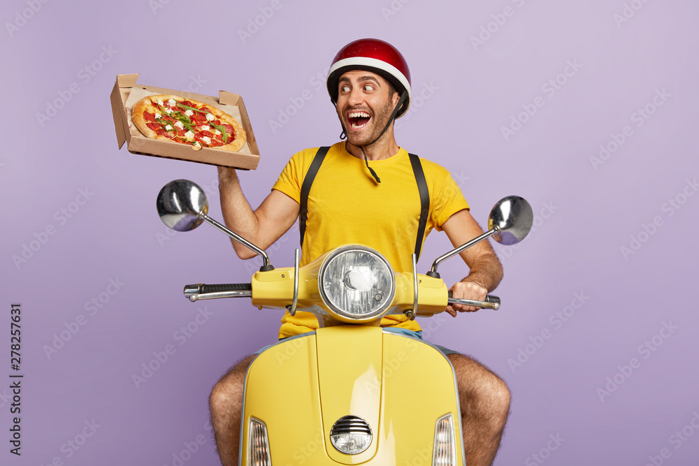 Wall mural fast food delivery concept. happy busy courier or deliveryman shows fresh baked pizza on box, poses 