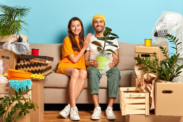 Delighted woman and man sit on sofa with household objects, buy luxury house to start new family...