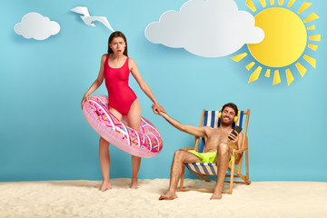 Lovely slim woman asks boyfriend to swim with her in sea, wears red bathing suit, poses inside of swimring. Relaxed man sits at beach chair, listens music via headphones in playlist, ignores wife