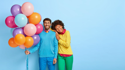 Horizontal shot of happy birthday man holds colorful balloons, Afro girl leans on his shoulder, wait for guests together, have broad smiles, isolated on blue wall with blank space. People and holiday