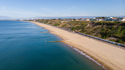 An aerial view of a majestic sandy beach with crystal blue water sea, groynes (breakwaters) and beach huts along a beautiful cliff with green vegetation under a blue and sunny sky