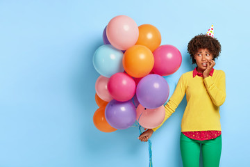 Fototapeta na wymiar Nervous puzzled woman feels shy and embarrassed in new company, gazes aside, bites finger nails, spends leisure time on party, holds bunch of colorful balloons wears festive paper hat, stylish apparel