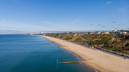 An aerial view of a majestic sandy beach with crystal blue water sea, groynes (breakwaters) and beach huts along a beautiful cliff with green vegetation under a blue and sunny sky