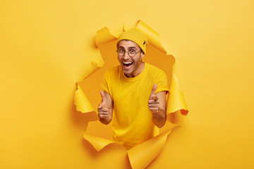 Joyful young optimistic man points fingers at you, has confident expression, poses through paper...