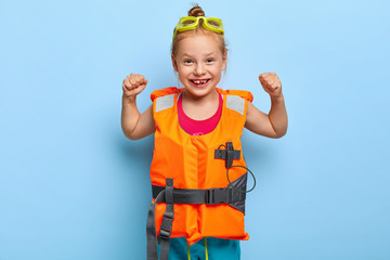 Joyful funny girl with red haired combed in bun, raises arms and shows muscles, wears protective...
