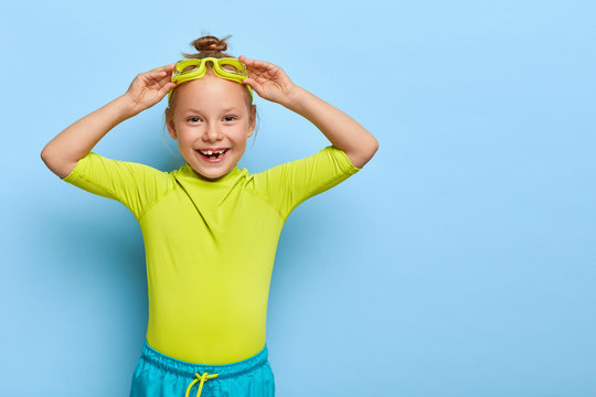 Pleasant looking cute small girl adjustes goggles, prepares for floating in pool, dressed in swimwear, has healthy body, poses indoor against blue wall. Copy space for text. Active rest for children