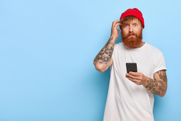 Embarrassed blue eyed man with ginger thick beard, scratches head, cannot understand how to use new...