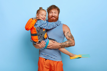 Affectionate father carries small red haired daughter in lifejacket and flippers, enjoy recreation...