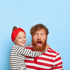 Photo of stupefied ginger father stares at camera, carries little daughter on hands, receives hug,...