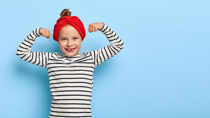 Strong happy small girl raises arms and shows biceps, has good physical shape, being active, wears...