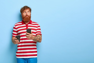 Pensive foxy guy focused aside, uses smartphone for browsing internet or chatting online, drinks...