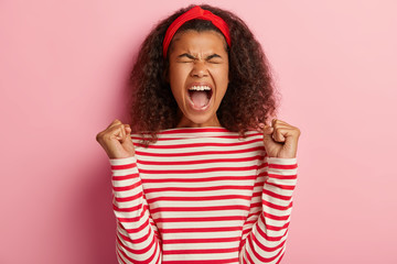 Finally success. Joyful curly young woman clenches fists with triumph, rejoices winning expensive prize, exclaims with joy, wears red headband and striped jumper, isolated on pink background