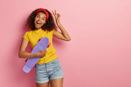 Enthusiastic girl with Afro haircut holds skateboard, makes peace gestue, has fun during leisure, tilts head, wears yellow t shirt and denim shorts, isolated on rosy wall. People, hobby, active rest