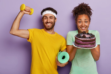 Cheerful motivated guy in yellow t shirt, raises arm with dumbbell, carries karemat, positive...