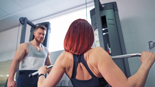 Close-up of female's back working on simulator indoors. Athletic woman is doing exercise while pulling the metal bar down at the fitness centre. Personal trainer man and a sportswoman.