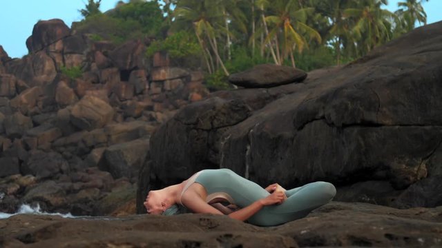 yoga practitioner changes position to easy fish asana on large stones against palm trees on rocky beach and sky extreme slow motion. Concept yoga meditation and spiritual practices