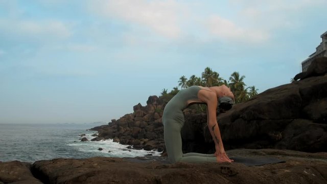 slim athletic lady stands in camel pose doing yoga training on rocky ocean beach against blue sky low angle shot extreme slow motion. Concept fitness sports yoga healthy lifestyle