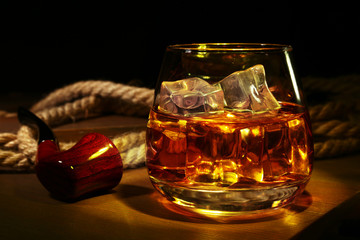 Glass with whiskey, ice cubes and smoking pipe on wooden background.