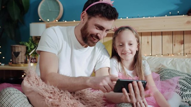 Portrait of pretty cheerful dad and his daughter watching videos and laughing of fun. Closeup little girl and her father play magic fairies dressed in pink having party at home.