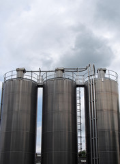 a group of three tall steel chemical storage tanks with connecting pipes and ladders against a blue cloudy sky