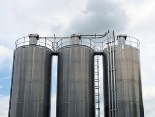 a group of three tall steel chemical storage tanks with connecting pipes and ladders against a blue cloudy sky