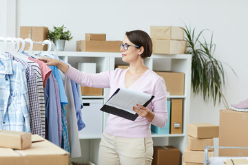 Young woman with document standing by rack and looking through new collection of casualwear