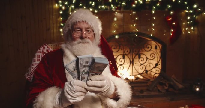 Authentic santa claus sitting in his rocker near fireplace and shaking packs of money in his hands - christmas spirit, holidays and celebrations concept 4k footage