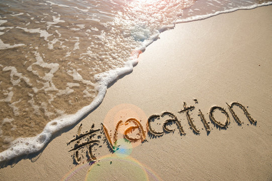 Modern travel message for the beach with a social media-friendly hashtag written with the word "vacation" in smooth sand with incoming wave