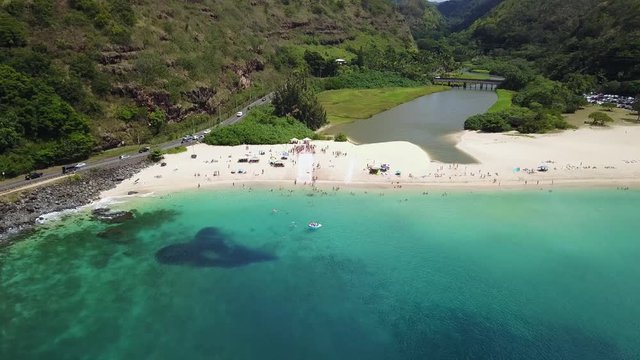 Drone Shot Flying Towards A Crowd Of People Surrounding A Slip N Slide On The Beach At Waimea Bay, Hawaii. People Can Be Seen Sliding Down The Slide Towards The Water.