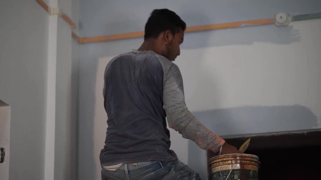 Indian man paints blue color on white wall above the door near the roof with paint brush, India