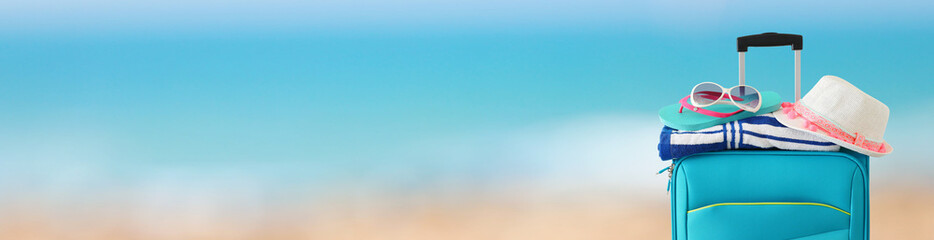 holidays. travel concept. blue suitcase with female hat, flip flips, sunglasses and beach towel in front of tropical background. banner © tomertu