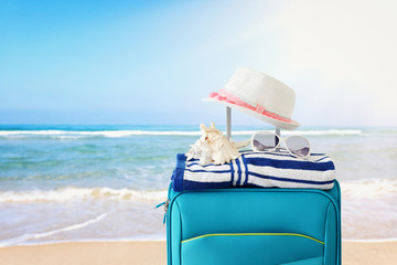 holidays. travel concept. blue suitcase with female hat, sea shell, sunglasses and beach towel in front of tropical background