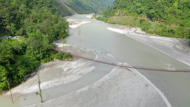 Aerial, tilt down, drone shot, towards a Lepcha clan man walking on a suspension bridge, over Tista river, on a sunny day, in Sikkim, India