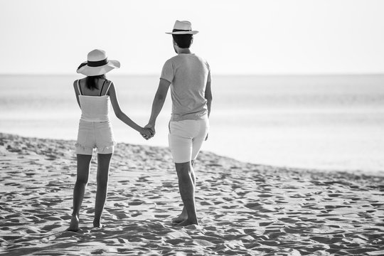 Back View Of Young Couple In Love  Looking The Future   And Holding Hand Together At Sea Beach  .happy Smiling Young Wedding With White Dress Romantic Moment  . Black And White Tone .hat . Rear