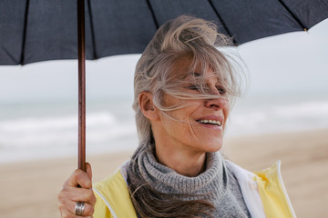Senior woman holding an umbrella in a rainy day on the beach.