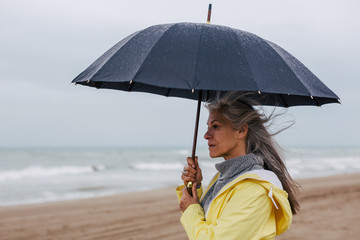 Senior woman holding an umbrella in a rainy day on the beach.