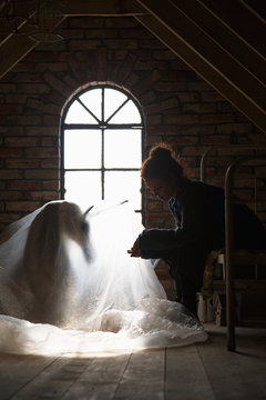 A girl with a unicorn doll sitting in an attic looking out the window