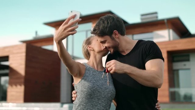 Happy couple making selfie photo with house keys outside residence.