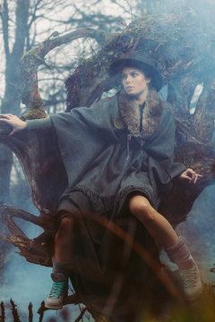 fashion model in loden outfit sitting on a overgrown root in foggy landscape