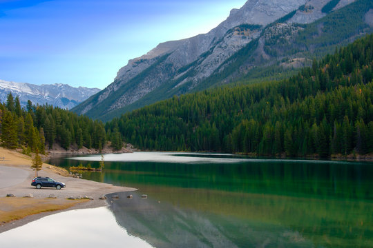 A Car Of Visitor Parking Next To Green Beatiful Lake With Huge Moutains Background. Concept Of Camping And Road Trip In Summer For Family And People Who Like Nature