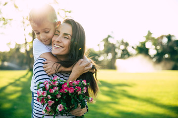 Present on mother's day. Little cute son makes surprise with flowers in hands for his happy and beautiful young mother outdoors in park