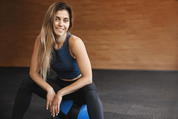 Close-up shot smiling sportswoman wearing activewear sit over wooden gym wall, workout with crossfit medicine ball, warm-up fitness exercise, look camera, talking health club instructor