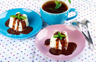 Cream pudding with chocolate sauce, mint and cup of tea on multicolored tablecloth.