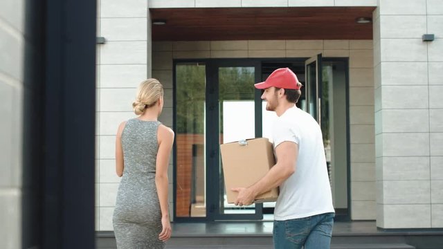 Young courier shipping parcel in box. Beautiful woman meeting postman near house